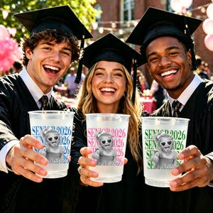 May include: Three graduates in black gowns and caps holding clear plastic cups. The cups have the text "SENIOR 2026" and a cartoon image of a child wearing sunglasses. The cups have different colored text: blue, pink, and green.