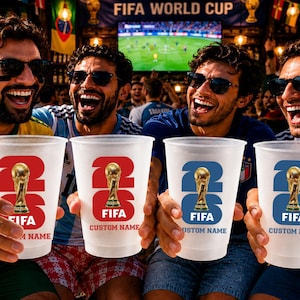 May include: Four people holding clear plastic cups with the FIFA logo and a golden trophy. The cups have the number 26 and the word FIFA printed on them. The background includes a FIFA World Cup banner and a television screen.