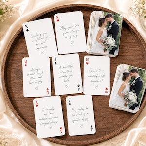 May include: A set of white playing cards featuring black and red text with wedding-themed messages, including cards with a photograph of a bride and groom. The cards are arranged on a round wooden tray with a gold pen and white floral accents.