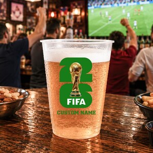 May include: A plastic pint glass filled with beer, displaying a green and gold FIFA logo. The glass has the text "FIFA" and "CUSTOM NAME". The background shows a bar with people watching a football match on a screen.