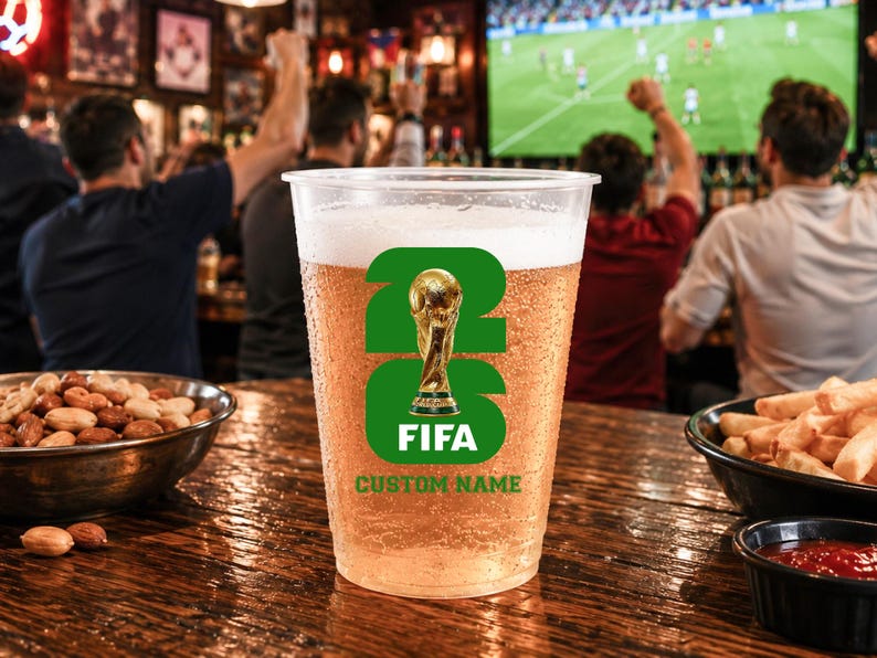 May include: A plastic pint glass filled with beer, displaying a green and gold FIFA logo. The glass has the text "FIFA" and "CUSTOM NAME". The background shows a bar with people watching a football match on a screen.