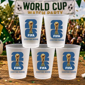 May include: A set of frosted plastic cups with a blue and gold FIFA World Cup design. Each cup features the number 23, a trophy graphic, and the text "FIFA CUSTOM NAME". A golden trophy and football are also visible.