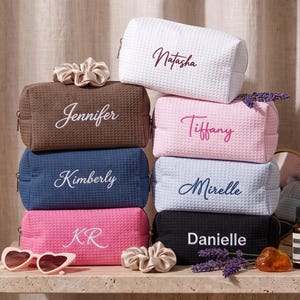 May include: A stack of rectangular waffle-textured cosmetic bags in white, brown, navy blue, pink, light blue, and black. Each bag features embroidered names including Natasha, Jennifer, Tiffany, Kimberly, Mirelle, KR, and Danielle, with scrunchies and sunglasses.