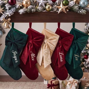 May include: Five velvet Christmas stockings hanging in a row, featuring large bows and embroidered names: Jasper, Alice, Charlie, Luna, and Leo. The stockings are colored in deep green, burgundy, and champagne gold, each with a matching fabric loop.