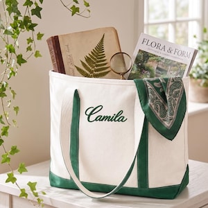 Peut inclure: Un sac fourre-tout en toile blanche avec des bordures et des anses vert foncé, portant le prénom Camila brodé en écriture cursive vert foncé. Le sac contient un livre avec une illustration de fougère, une loupe et un magazine Flora & Form.