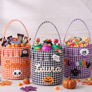 May include: Three fabric Halloween buckets with gingham patterns in orange, black, and purple. Each bucket features a handle, a pleated top edge, and a personalized name in felt lettering: Sarah, Laura, and Timothy. They are filled with assorted candy.