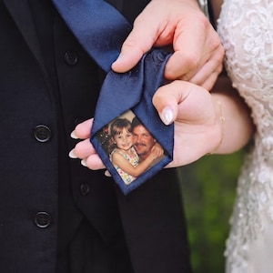 Puede incluir: Una corbata azul marino oscuro con un sutil patrón de cachemira y un parche fotográfico rectangular personalizado cosido en la parte posterior. La foto muestra a un niño de pelo castaño con una blusa floral abrazando a un adulto con bigote.