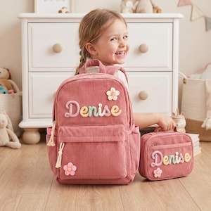 May include: A dusty rose corduroy backpack and matching lunch bag set, both featuring the name Denise in colorful, textured, raised lettering. Each item is decorated with small, soft, fabric flower appliqués and includes cream-colored zipper pulls.