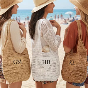 Pode incluir: Três bolsas de crochê com alças de ombro, cada uma com um remendo triangular com iniciais bordadas. Duas bolsas são bege e uma é branca. As bolsas exibem as letras GM, HB e KL, com uma concha visível dentro da bolsa branca.