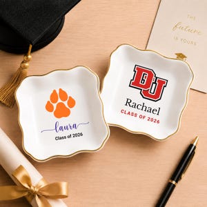 May include: Two white ceramic trinket dishes with gold rims. One features an orange paw print and the name "laura" with "Class of 2026". The other has a red and black logo, the name "Rachael", and "Class of 2026". A graduation cap and diploma are in the background.