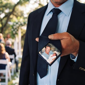 May include: A dark navy blue necktie featuring a hidden, diamond-shaped photograph patch sewn onto the underside of the wide end. The photograph depicts a person holding a newborn baby wrapped in a white blanket, visible when the tie is lifted.