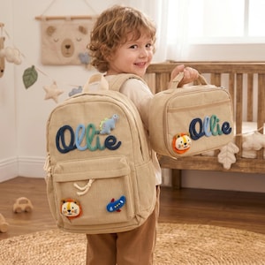 May include: A beige corduroy backpack and a matching lunch bag, both featuring the name Ollie embroidered in blue and green yarn. The backpack includes a small plush dinosaur, a lion face, and an airplane patch. The lunch bag features a lion face.