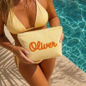 K&ouml;nnte beinhalten: Eine gelbe Cord-Tasche mit dem Namen "Oliver" in Orange gestickt. Die Tasche wird vor einer Person gehalten, die einen gelben Bikini tr&auml;gt. Der Hintergrund zeigt einen Swimmingpool.
