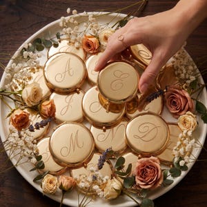 May include: A collection of round, gold-toned metal compact mirrors, each engraved with a unique cursive letter. The mirrors are arranged on a white plate, interspersed with small peach-colored roses, sprigs of baby's breath, and lavender.