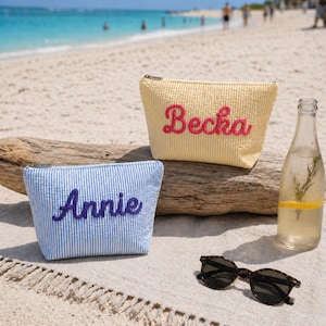 K&ouml;nnte beinhalten: Zwei personalisierte Kosmetiktaschen am Strand. Eine ist gelb mit "Becka" in rosa Schrift, die andere blau und wei&szlig; gestreift mit "Annie" in lila Schrift. Eine Wasserflasche mit Zitrone und Rosmarin und eine Sonnenbrille sind ebenfalls am Strand.