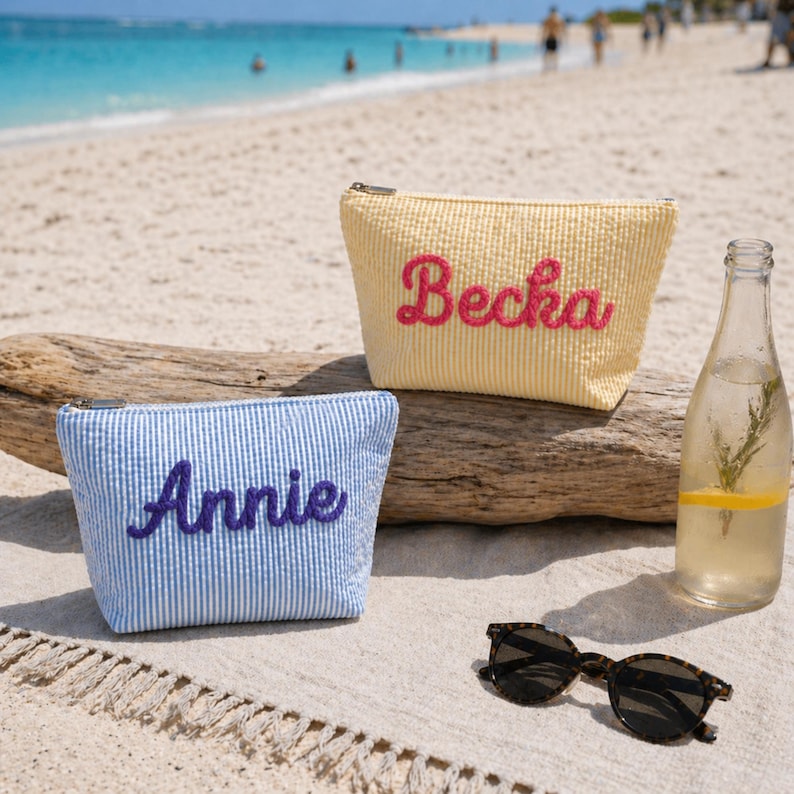 K&ouml;nnte beinhalten: Zwei personalisierte Kosmetiktaschen am Strand. Eine ist gelb mit "Becka" in rosa Schrift, die andere blau und wei&szlig; gestreift mit "Annie" in lila Schrift. Eine Wasserflasche mit Zitrone und Rosmarin und eine Sonnenbrille sind ebenfalls am Strand.