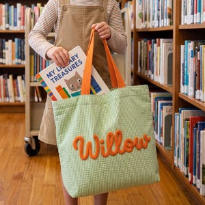 Op de afbeelding: Een lichtgroene seersucker tote bag met oranje stoffen hengsels en de naam Willow geborduurd in oranje garen op de voorzijde. De tas wordt vastgehouden naast een boek met de titel My Library Treasures met een kattenillustratie op de kaft.
