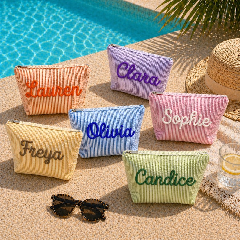 K&ouml;nnte beinhalten: Sechs personalisierte Kosmetiktaschen in Orange, Lila, Rosa, Blau, Gelb und Gr&uuml;n, jeweils mit einem gestickten Namen. Eine Sonnenbrille und ein Strohhut sind ebenfalls zu sehen. Die Taschen sind gestreift.