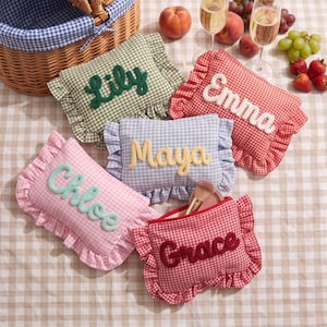 May include: Five rectangular zippered pouches with ruffled edges and gingham patterns in green, red, light blue, and pink. Each pouch features a name embroidered in raised, contrasting yarn: Lily, Emma, Maya, Chloe, and Grace. One pouch is open.