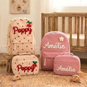 May include: Two sets of corduroy backpacks and lunch bags. One set is beige with a red strawberry pattern and the name Poppy in red script. The other set is dusty pink with a white flower appliqué and the name Amélie in white script.