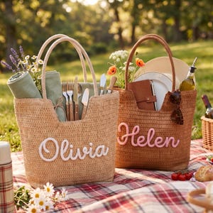 May include: Two woven straw tote bags with open tops and dual handles. The left bag features the white script name Olivia, and the right bag features the pink script name Helen. Both bags are filled with picnic supplies, including napkins, cutlery, plates, and flowers.