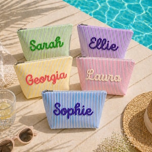 K&ouml;nnte beinhalten: F&uuml;nf gestreifte Kosmetiktaschen in Gr&uuml;n, Lila, Gelb, Rosa und Blau. Jede Tasche hat einen anderen Namen aufgestickt: Sarah, Ellie, Georgia, Laura und Sophie. Die Taschen sind mit Rei&szlig;verschl&uuml;ssen versehen und liegen auf einer Steinoberfl&auml;che in der N&auml;he eines Pools.