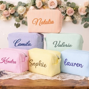 May include: A collection of six rectangular, ribbed fabric cosmetic bags in pastel colors, each featuring a personalized name embroidered in a cursive font. The bags are arranged in a pyramid stack, showcasing shades of peach, purple, mint, pink, yellow, and blue.