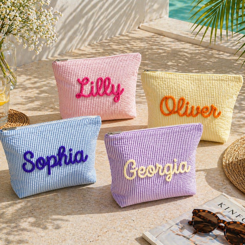 K&ouml;nnte beinhalten: Vier Kosmetiktaschen aus Seersucker in Rosa, Gelb, Blau und Lila, jeweils mit einem Namen bestickt: Lilly, Oliver, Sophia und Georgia. Die Taschen haben einen Rei&szlig;verschluss. Das Bild wurde im Freien in der N&auml;he eines Pools aufgenommen.