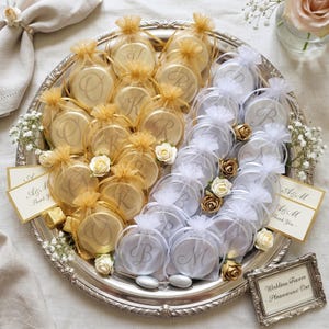 May include: A silver platter holds round wedding favors in gold and white organza bags, each featuring a monogram. The display includes small white and gold artificial roses, sprigs of baby's breath, and cards reading A&M Thank You, plus a sign reading Wedding Favors Please take one.