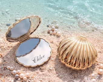 May include: Two gold-toned, shell-shaped compact mirrors. One mirror is open, revealing a reflective surface with the name Chloe printed in black script. The other mirror is closed, displaying a ribbed, metallic exterior shell design.