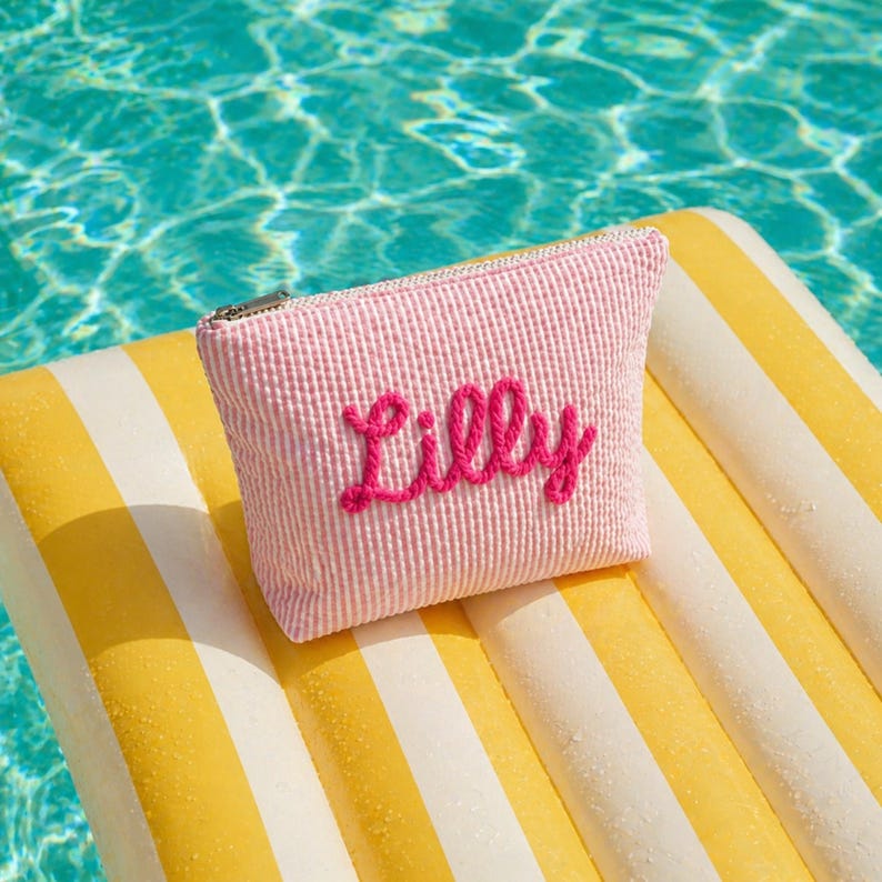 K&ouml;nnte beinhalten: Ein rosa Kord-Rei&szlig;verschlussetui mit dem Namen "Lilly" in Schreibschrift gestickt. Das Etui liegt auf einer gelb-wei&szlig; gestreiften Pool-Schwimminsel, mit einem t&uuml;rkisfarbenen Pool im Hintergrund.