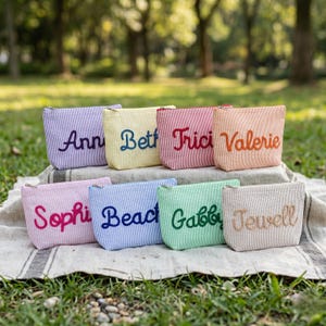 May include: Eight rectangular zippered pouches made of seersucker fabric in various pastel colors, each featuring a name embroidered in a cursive font. The pouches are arranged in two rows, displaying names including Ann, Beth, Tricia, Valerie, Sophia, Beach, Gabby, and Jewell.
