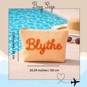 K&ouml;nnte beinhalten: Eine orangefarbene, strukturierte Kosmetiktasche mit dem Namen "Blythe", der mit orangefarbenem Faden gestickt ist. Die Tasche ist 18 cm hoch und 26 cm breit. Ein Strohhut und eine Sonnenbrille sind im Hintergrund.