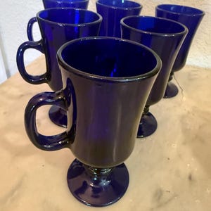 Op de afbeelding: Een set van zes kobaltblauwe glazen mokken, elk met een gladde, ronde kom, een stevig C-vormig handvat en een voetstuk. Het diepblauwe glas is doorschijnend en reflecteert het licht over de gebogen oppervlakken van de bijpassende drinkglazen.