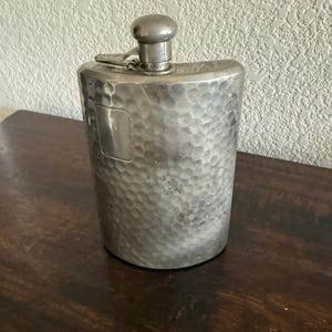 Puede incluir: Una petaca de metal plateado con una textura martillada en todo su cuerpo curvo. La petaca incluye una placa rectangular lisa y vacía en la parte superior izquierda y un tapón de rosca redondeado unido a un brazo metálico articulado.