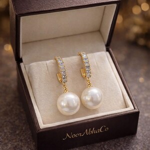 Puede incluir: Un par de pendientes de aro dorados con cristales transparentes y grandes perlas blancas. Los pendientes se muestran en una caja de joyería marrón con el texto "NoorAbhaCo" en la parte inferior.
