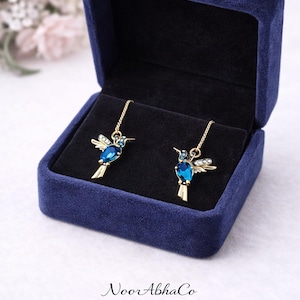 May include: Gold-coloured hummingbird earrings with blue gemstone bodies and crystal accents, presented in a navy blue velvet jewellery box. The earrings hang from fine chains. The open box reveals a black interior.