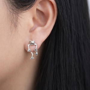 May include: Silver frog earrings. The earrings feature a frog design, with the frog's body on the ear and its legs hanging down. The earrings are detailed and have a whimsical design. The earrings are worn on a person's ear.