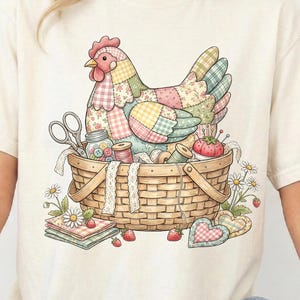 May include: Cream-colored t-shirt featuring a colorful patchwork chicken design. The chicken sits in a woven basket filled with sewing supplies, including scissors, spools of thread, and a pincushion. Daisies and strawberries surround the basket.