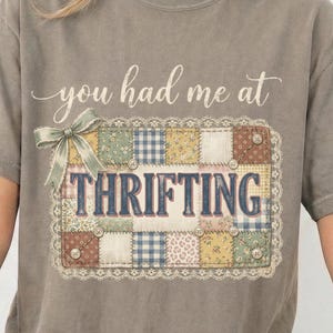 May include: A gray t-shirt with the phrase "you had me at THRIFTING" printed on the front. The text is above a patchwork design with a bow. The patchwork design includes various patterns and colors.