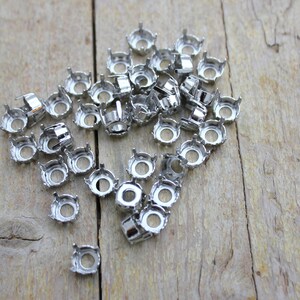 Puede incluir: Primer plano de un montón de engastes metálicos plateados para diamantes de imitación. Los engastes son redondos con un borde con puntas y un agujero en el centro.