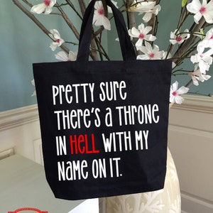 May include: Black tote bag with white text that reads "Pretty sure There's a Throne IN HELL WITH MY Name ON IT." The word "HELL" is in red. The bag is hanging in front of a vase with white flowers.