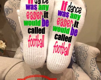 May include: A pair of white socks with the text "If dance was any easier, it would be called football" in various colors. The socks are resting on a patterned chair.