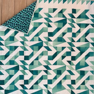 Puede incluir: Un edredón hecho a mano con un diseño geométrico en tonos turquesa y blanco. El edredón presenta un patrón repetitivo de triángulos y cuadrados, con un patrón floral turquesa contrastante en el reverso. El edredón está doblado, revelando ambos lados.