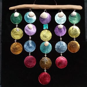 May include: A vibrant wind chime with iridescent, circular shells in shades of green, purple, and gold. Each shell is decorated with a seashell and linked with clear beads. The chime is suspended from a natural wooden branch.