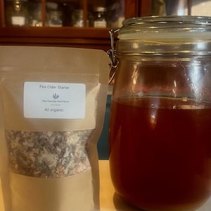 May include: A glass jar with a metal clasp and a brown liquid next to a brown paper bag. The bag has a white label that reads "Fire Cider Starter" and "All organic". The jar is partially filled with a reddish-brown liquid.