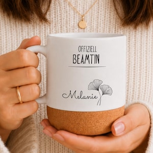 Könnte beinhalten: Ein weißer Keramikbecher mit Korkboden, der auf der Vorderseite den schwarzen Aufdruck "OFFIZIELL BEAMTIN" über einer Linie sowie den Namen "Melanie" in Schreibschrift neben einer schwarzen Strichzeichnung eines Ginkgo-Zweiges zeigt.