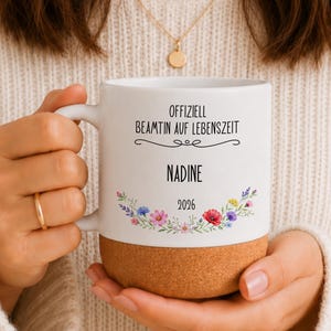 Könnte beinhalten: Ein weißer Keramikbecher mit Korkboden, bedruckt mit dem schwarzen Text "OFFIZIELL BEAMTIN AUF LEBENSZEIT", dem Namen "NADINE", der Jahreszahl "2026" und einer bunten Blumenillustration in den Farben Rosa, Blau, Violett und Gelb.