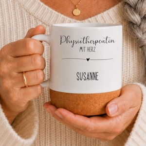 Könnte beinhalten: Ein weißer Keramikbecher mit Korkboden, der den schwarzen Aufdruck Physiotherapeutin MIT HERZ und den Namen SUSANNE trägt, getrennt durch eine horizontale Linie mit einem kleinen Herzsymbol in der Mitte, gehalten von zwei Händen.