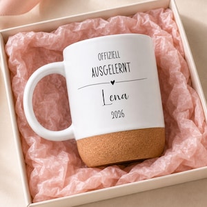 Könnte beinhalten: Ein weißer Keramikbecher mit Korkboden, bedruckt mit dem schwarzen Text "OFFIZIELL AUSGELERNT", einem kleinen Herzsymbol, dem Namen "Lena" und der Jahreszahl "2026". Der Becher liegt in einer quadratischen Geschenkbox mit hellrosa Seidenpapier.